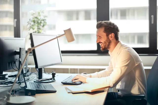 A man smiling while working at an office desk with a computer and natural daylight streaming in through large windows. Running email software with a durable record for every campaign